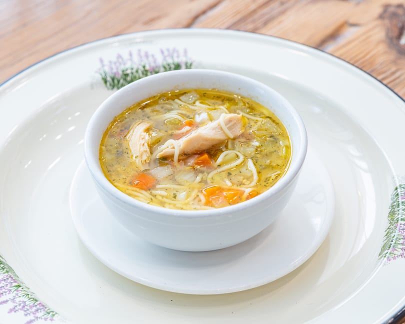 Chicken noodle soup with carrots and herbs in a white bowl on a decorative plate.