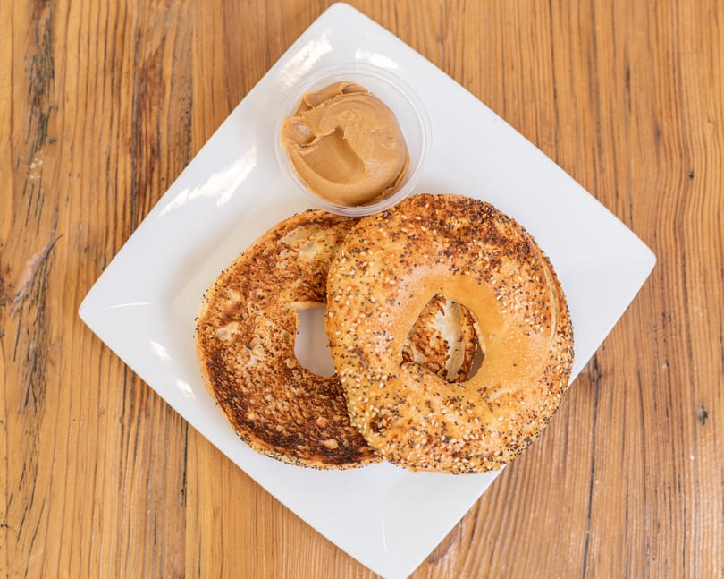 Two toasted bagel halves, one everything, with a side of peanut butter on a white plate.