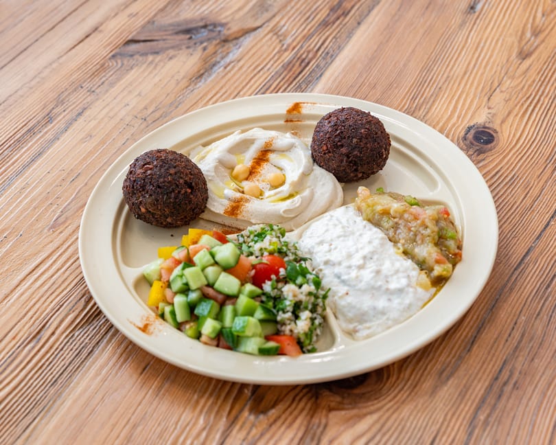 A beige plate with falafel, hummus, tabbouleh, baba ghanoush, and a white dip on a wooden table.