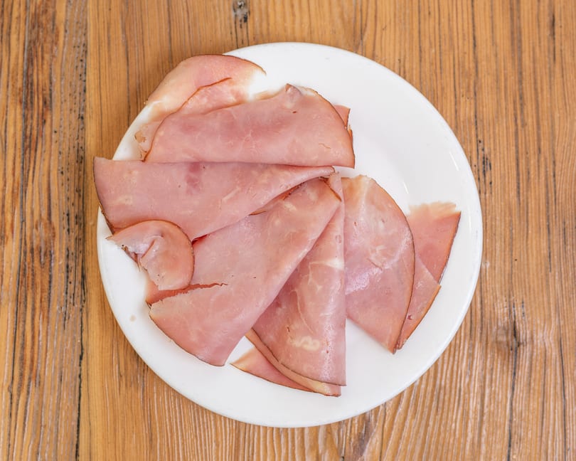 A white plate filled with folded slices of pink deli ham on a wooden table.