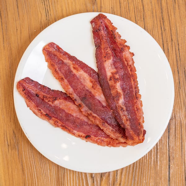 Three crispy bacon strips on a white plate, resting on a wooden table.