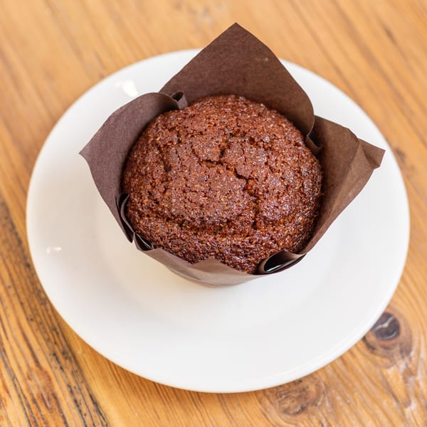 Chocolate muffin in a brown paper wrapper on a white plate, resting on a wooden table.