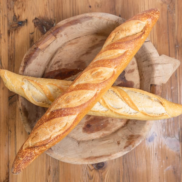 Two golden-brown baguettes rest on a rustic wooden platter on a wooden table.