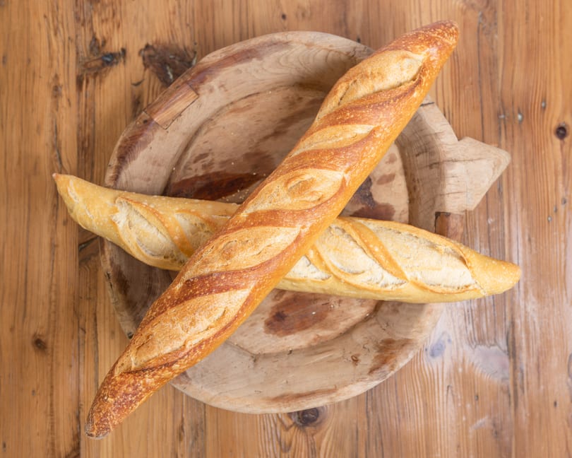 Two golden-brown baguettes rest on a rustic wooden platter on a wooden table.