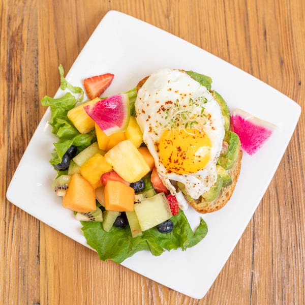 Avocado toast with a fried egg and a side of colorful fruit salad on a white plate.