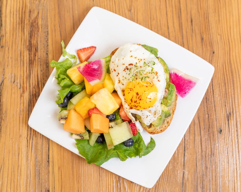 Avocado toast with a fried egg and a side of colorful fruit salad on a white plate.