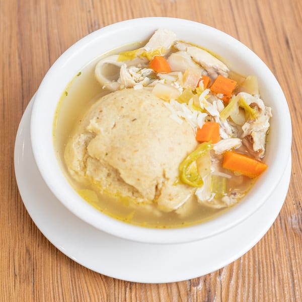 White bowl of chicken matzo ball soup with carrots and celery on a wooden table.