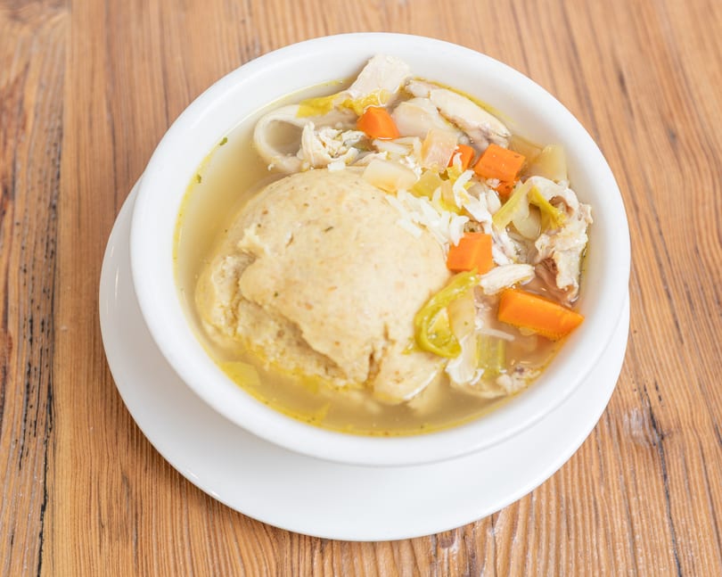 White bowl of chicken matzo ball soup with carrots and celery on a wooden table.