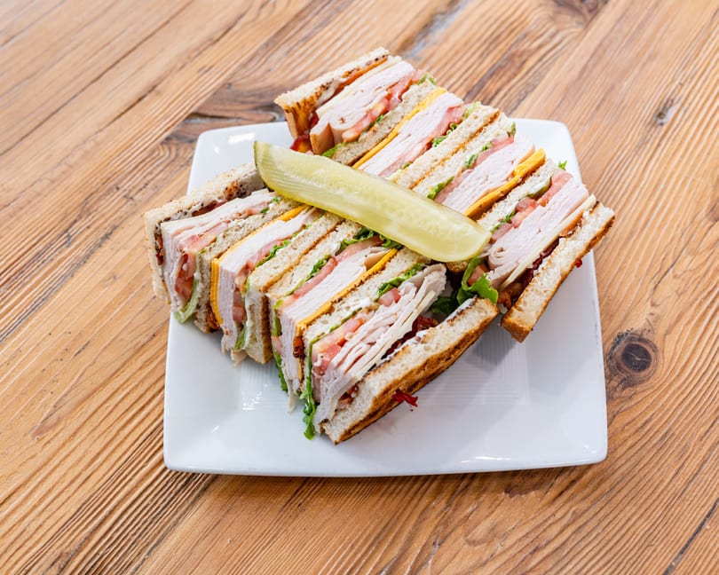 Club sandwich halves with turkey, cheese, bacon, lettuce, tomato, and a pickle spear.
