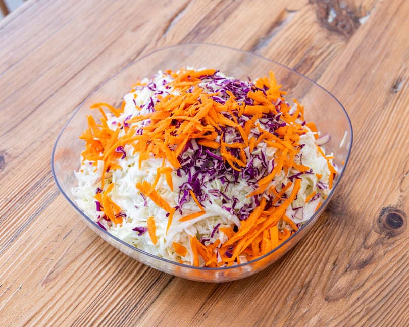 A clear bowl filled with shredded white cabbage, purple cabbage, and orange carrots on a wooden table.