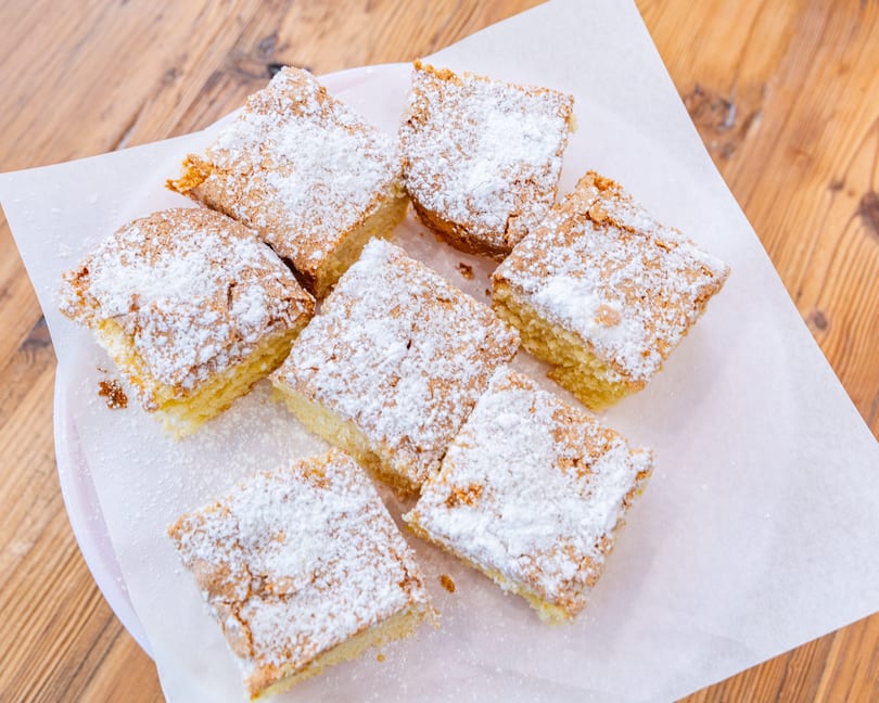 Seven square pieces of cake with powdered sugar on white parchment paper on a wooden table.